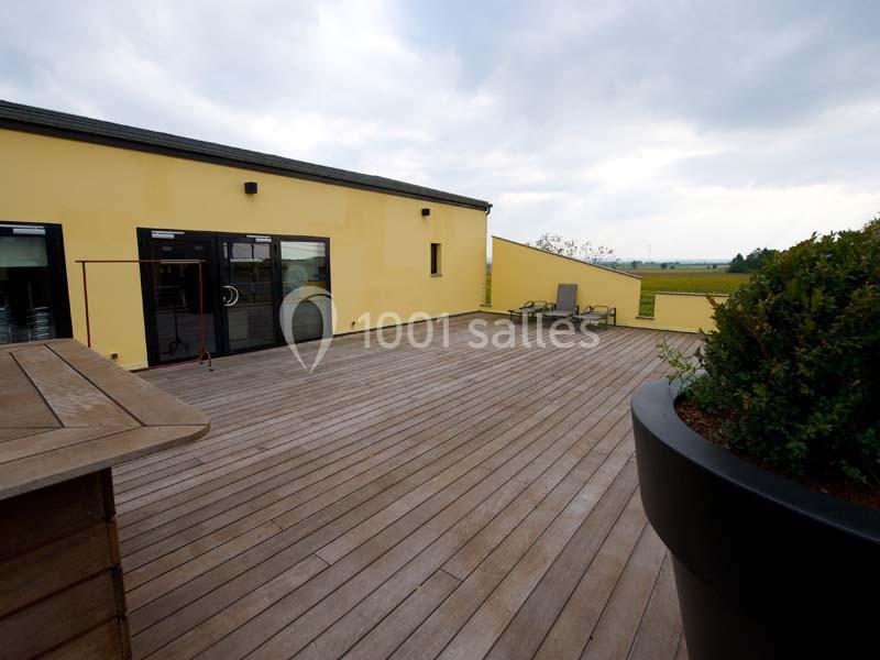 Terrasse en bois avec des chaises longues, bordée par un bâtiment jaune et offrant une vue dégagée sur la campagne.