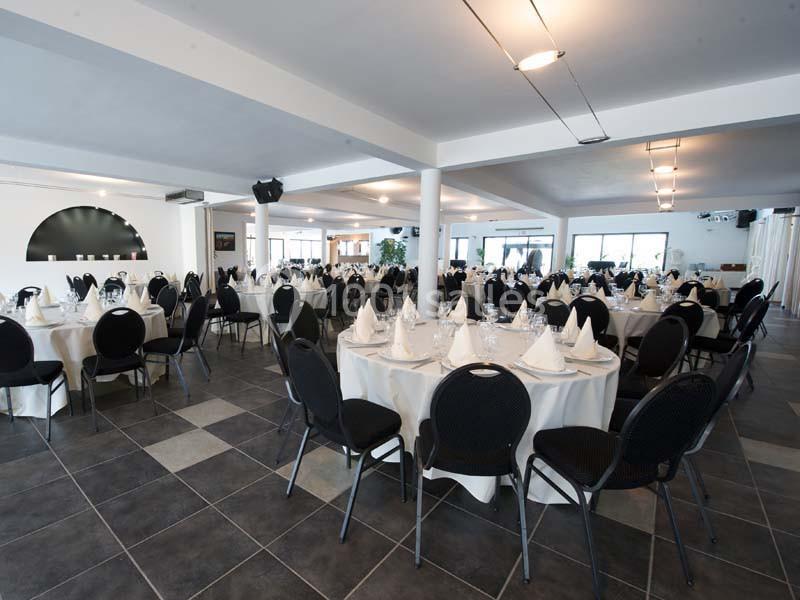 Salle de réception lumineuse avec tables rondes dressées de nappes blanches et chaises noires.