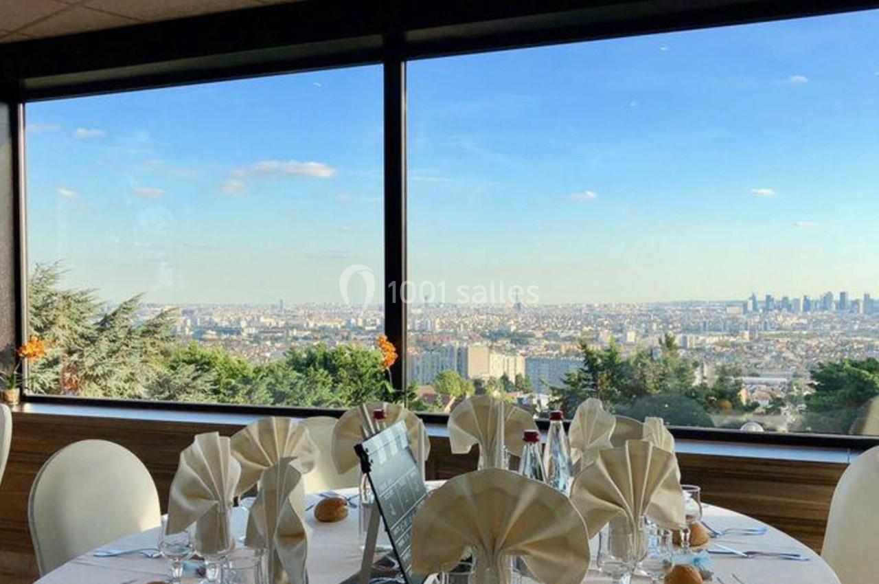 Vue panoramique sur une ville depuis une salle de réception avec des tables dressées et des grandes baies vitrées.