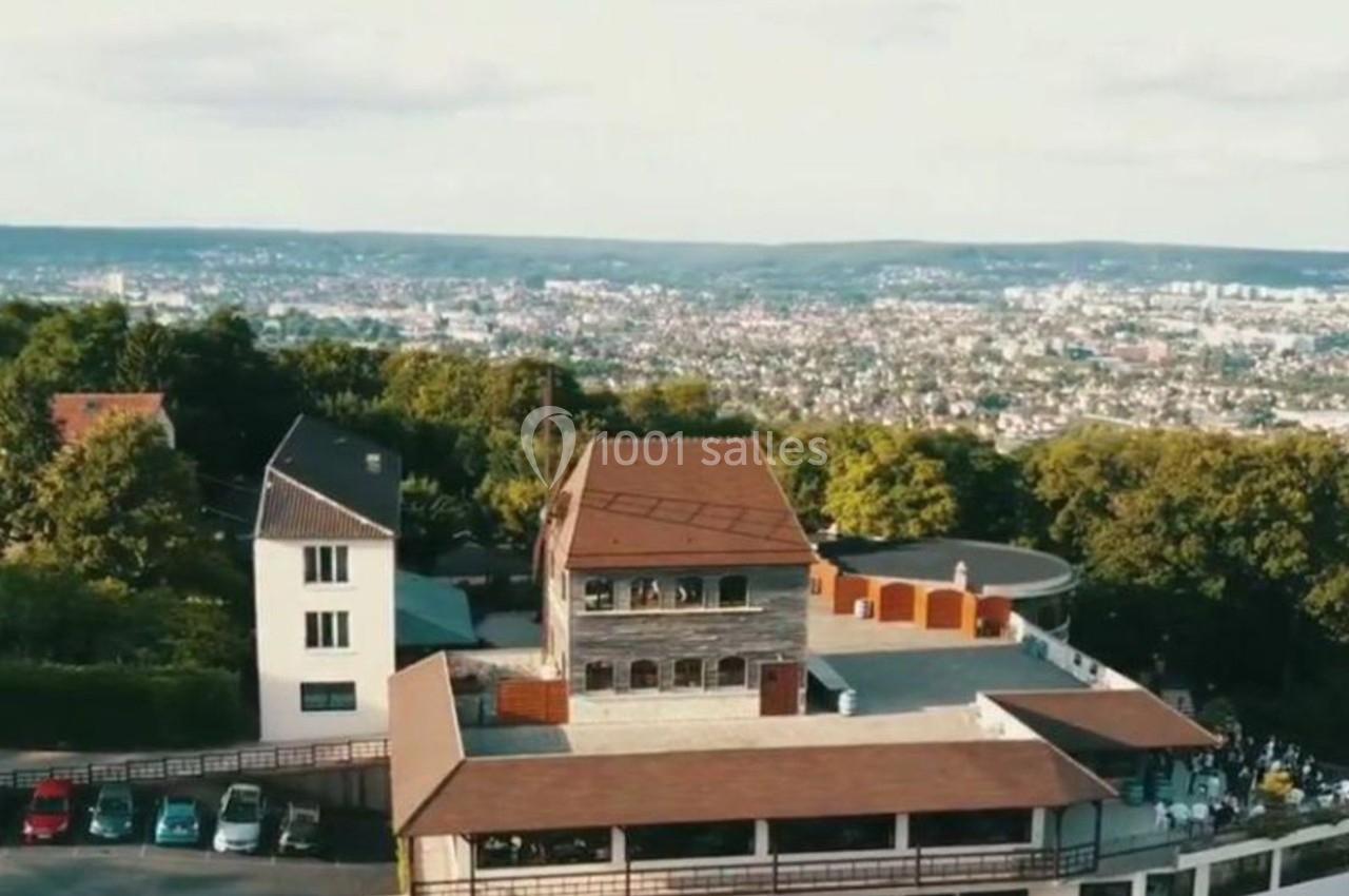 Vue aérienne d'un bâtiment en hauteur entouré de verdure, avec une ville visible à l'arrière-plan.