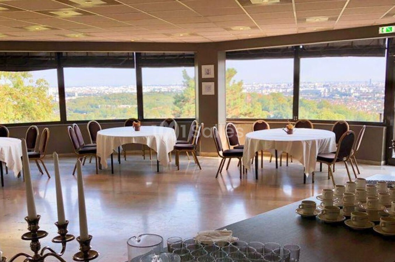 Salle lumineuse avec grandes baies vitrées, tables rondes dressées et vue panoramique sur un paysage urbain et verdoyant.