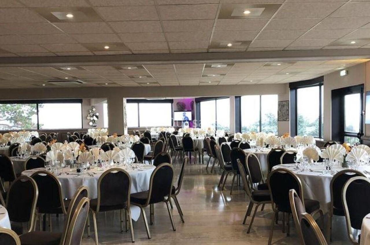 Salle de réception lumineuse avec des tables rondes dressées, nappes blanches et chaises noires.