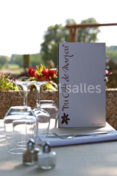 Menu posé sur une table dressée en extérieur, entourée de verres, fleurs et paysage verdoyant en arrière-plan.