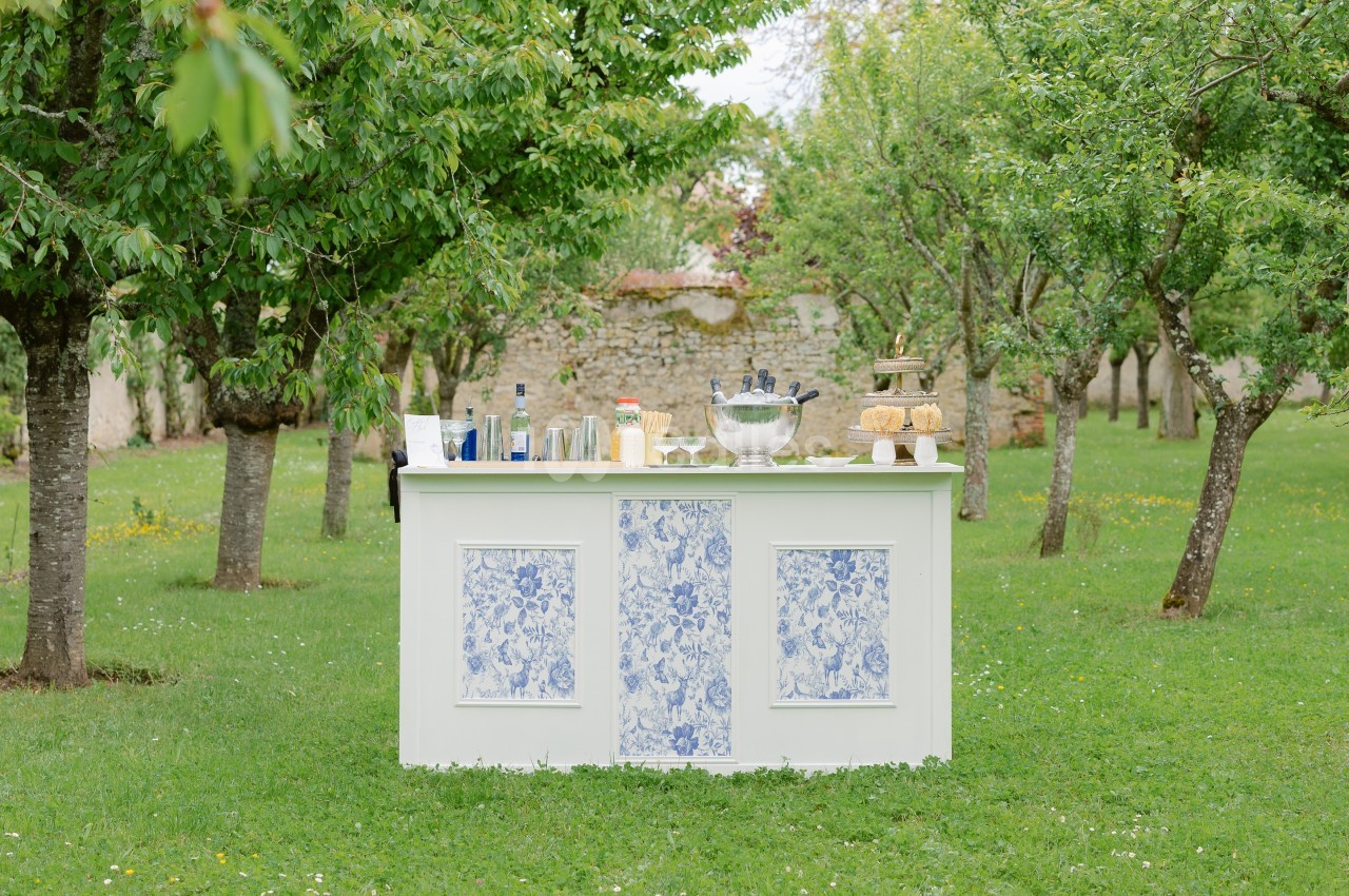 Bar extérieur blanc décoré de motifs bleus, installé dans un jardin verdoyant avec des arbres et des boissons disposées…