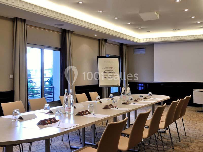 Salle de réunion lumineuse avec table ovale, chaises beiges, écran de projection et bouteilles d'eau disposées.