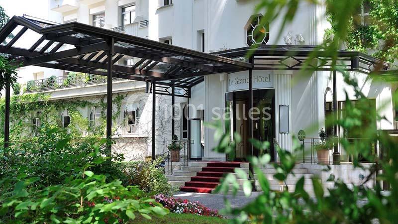 Entrée d'un hôtel avec une verrière, des escaliers bordés de tapis rouge et des plantes environnantes.