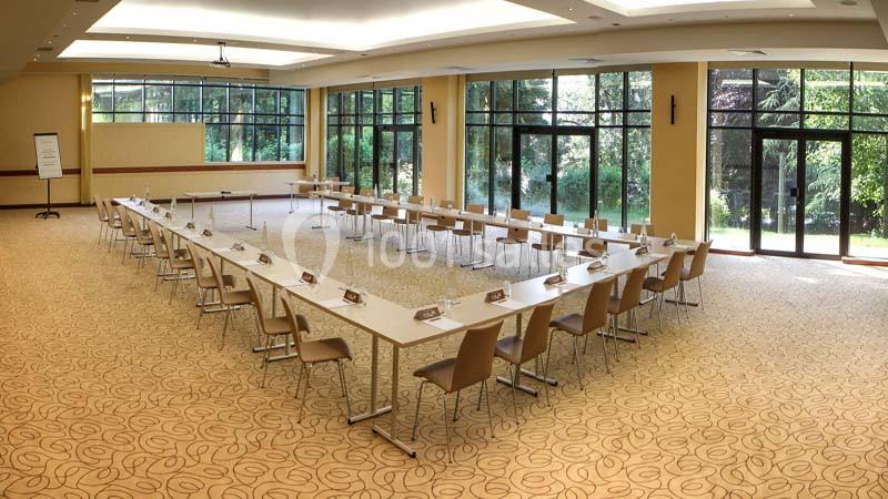 Salle de réunion lumineuse avec tables disposées en U, chaises en bois clair et grandes baies vitrées donnant sur un jardin.