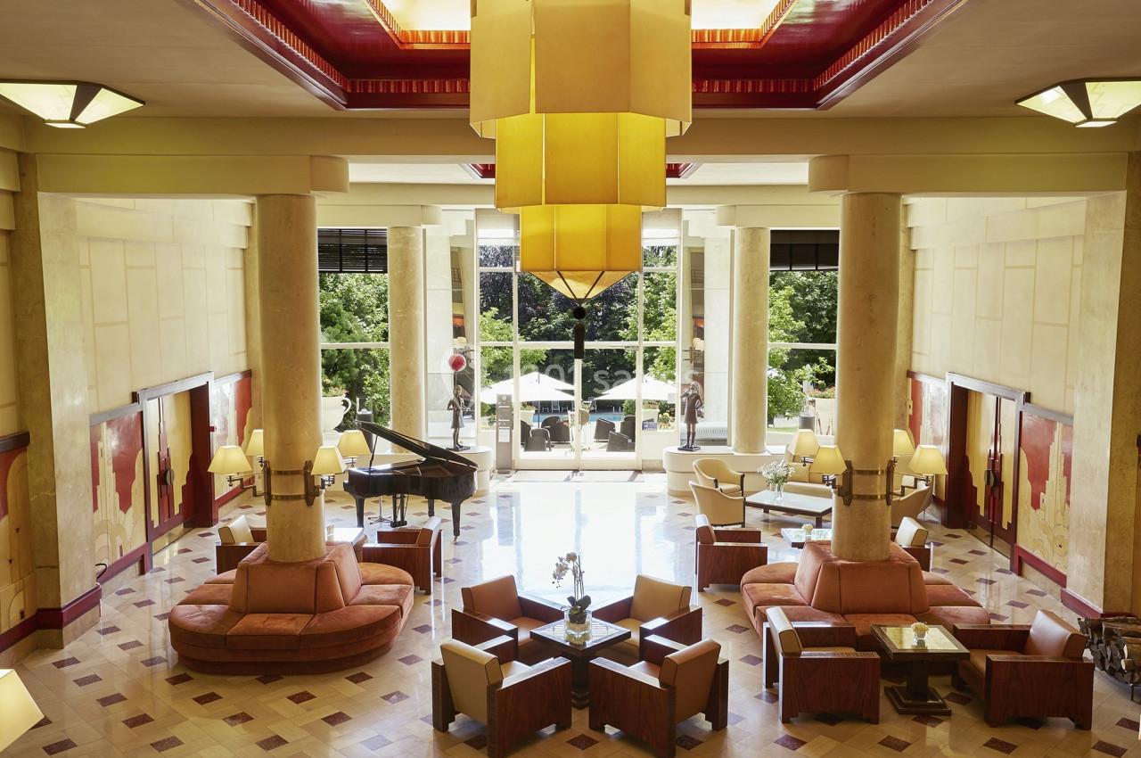 Hall spacieux avec colonnes, mobilier élégant, piano à queue et grandes baies vitrées donnant sur une terrasse verdoyante.