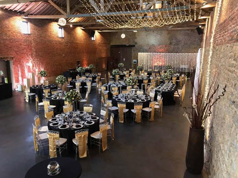 Salle de réception décorée avec des tables rondes, nappes noires, chaises dorées et guirlandes lumineuses au plafond.