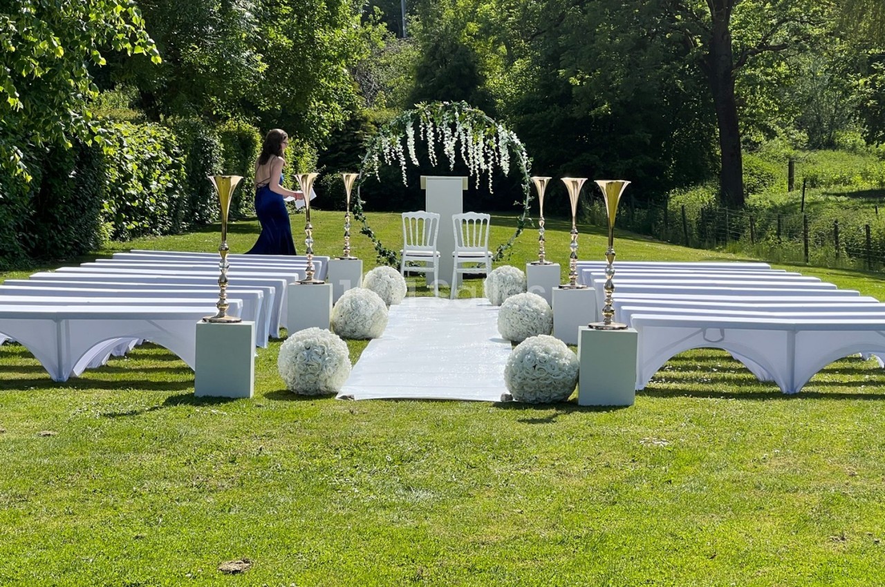 Allée centrale décorée pour une cérémonie en extérieur, avec chaises blanches, arches fleuries et pelouse verdoyante.