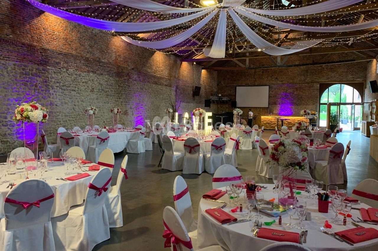 Salle de réception décorée pour un mariage, avec tables dressées, nappes blanches, rubans roses et éclairage tamisé.