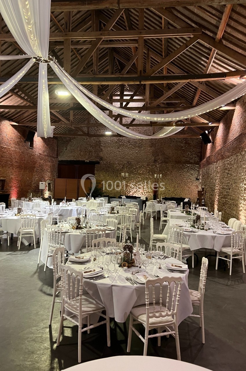 Salle de réception en pierre avec tables rondes dressées, nappes blanches et décorations suspendues au plafond.
