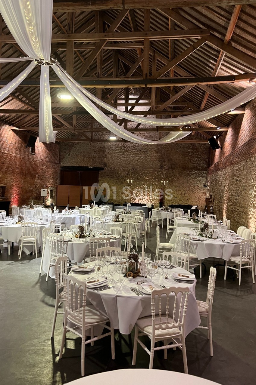 Salle de réception en pierre avec tables rondes dressées, nappes blanches et décorations suspendues au plafond.