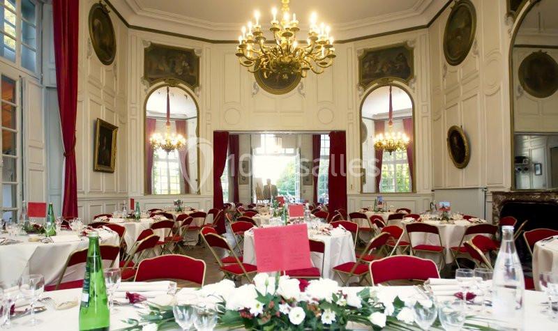 Salle de réception élégante avec tables dressées, chaises rouges, lustre central et grandes fenêtres.