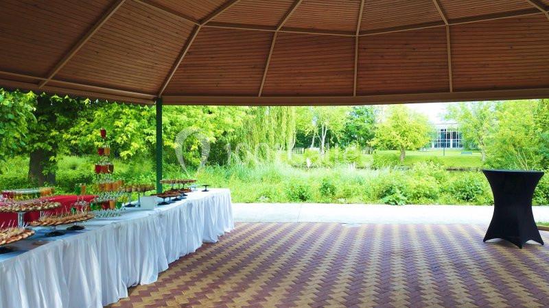 Table dressée sous un kiosque en bois avec vue sur un jardin verdoyant et un bâtiment au loin.