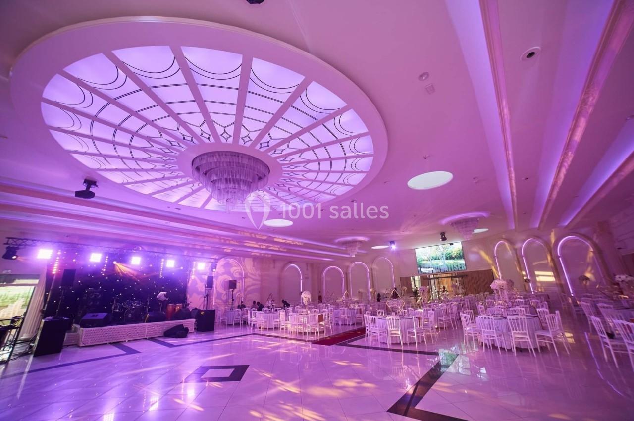 Salle de réception spacieuse avec plafond lumineux, tables décorées et scène éclairée pour un événement festif.