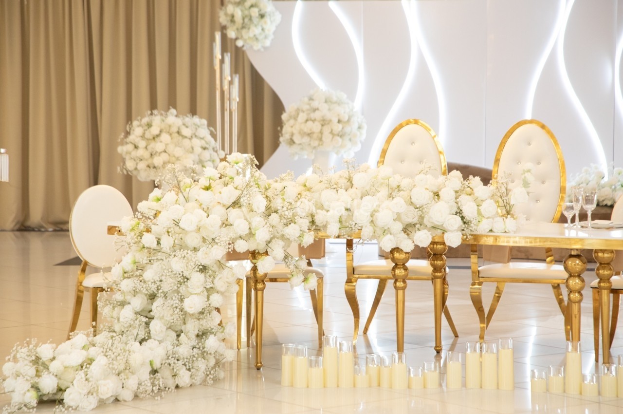 Table décorée de fleurs blanches, chaises dorées et bougies alignées dans une salle lumineuse avec rideaux beiges.