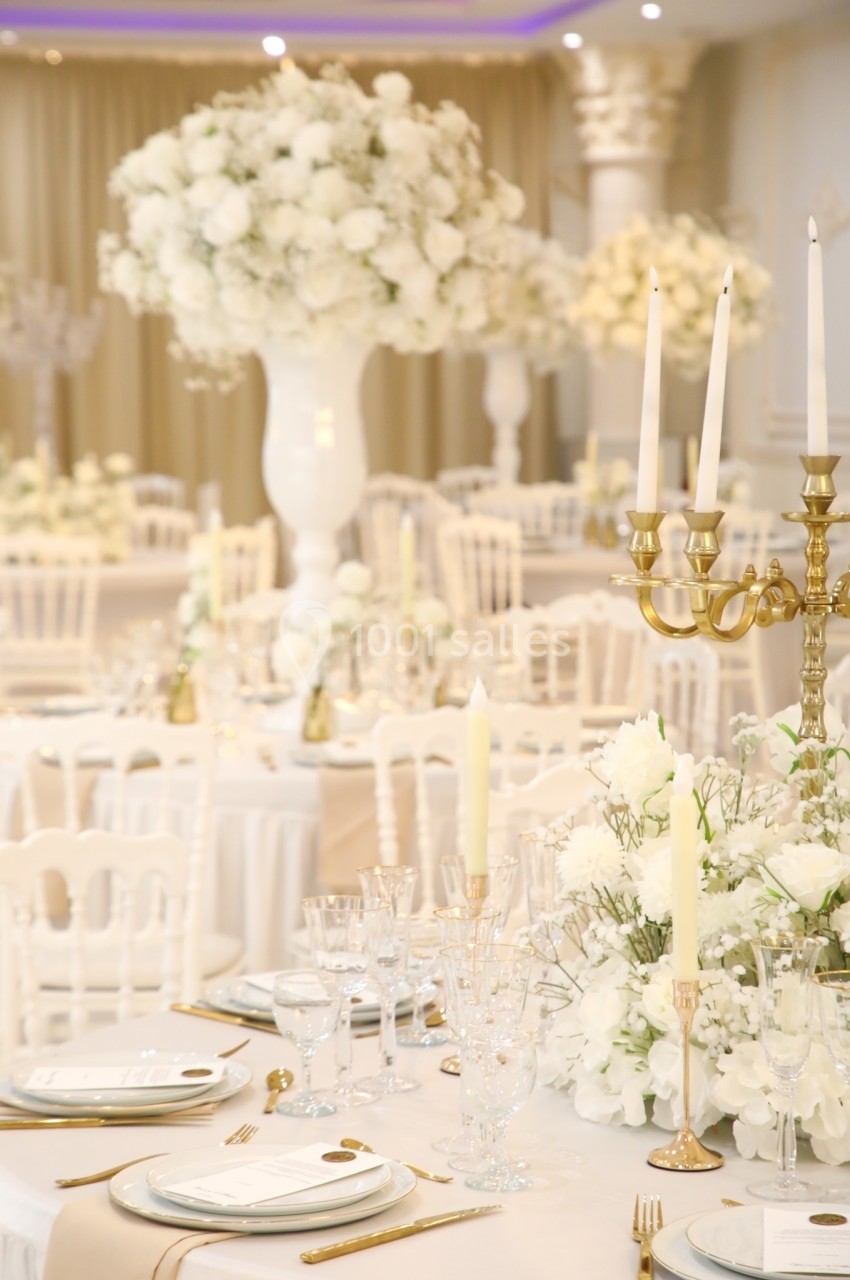 Salle de réception élégante décorée de fleurs blanches, chandeliers dorés et tables dressées avec vaisselle raffinée.