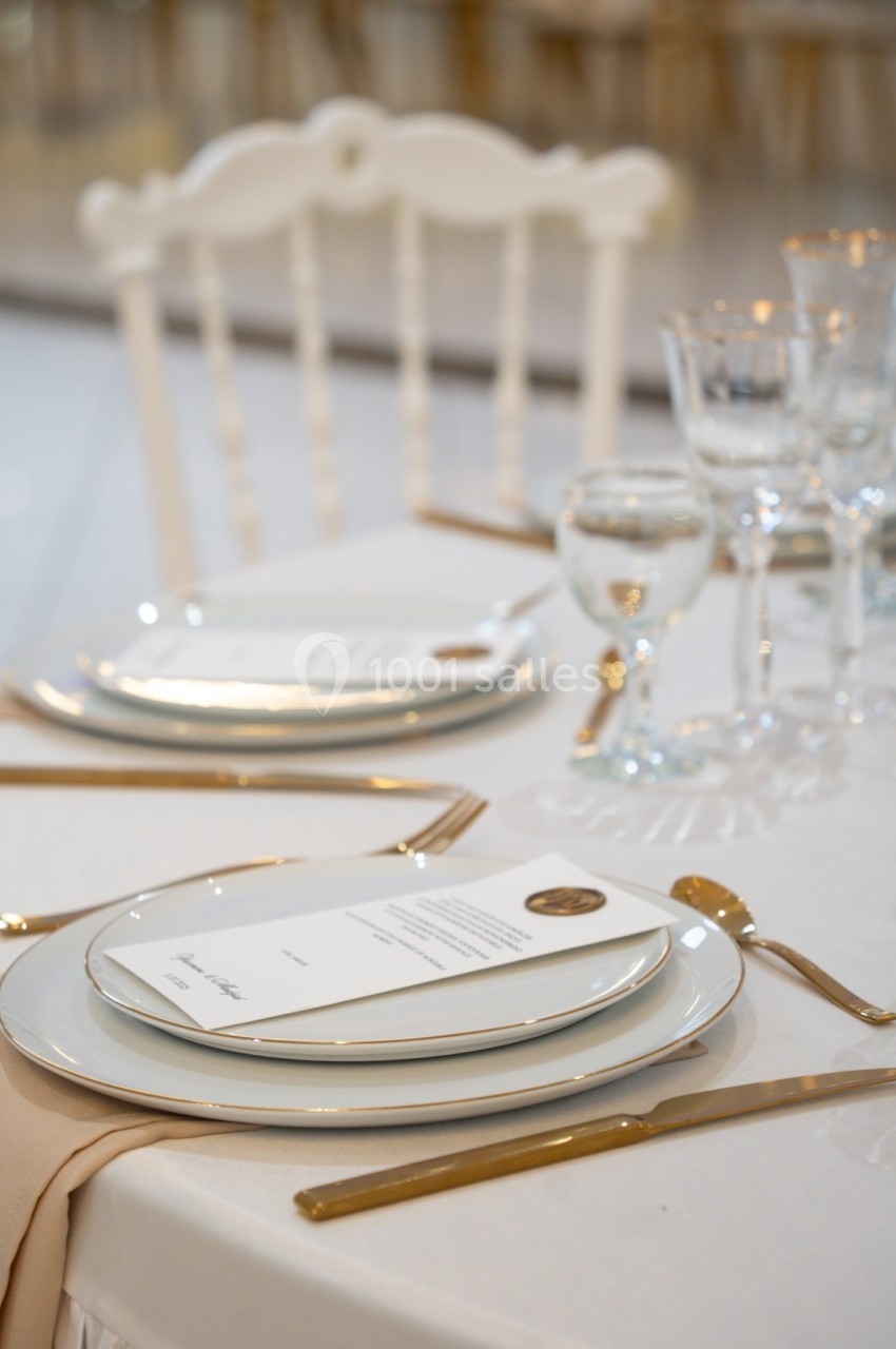 Table dressée avec vaisselle blanche, couverts dorés, serviette beige et menus posés sur les assiettes.