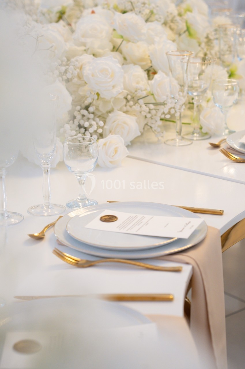 Table élégamment dressée avec des couverts dorés, assiettes blanches, menus et décoration florale blanche.