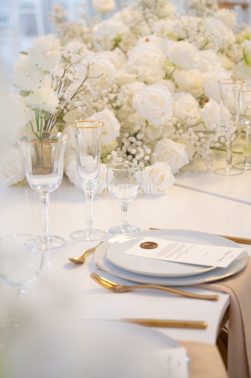 Table élégamment dressée avec vaisselle raffinée, couverts dorés et centre de table floral blanc.