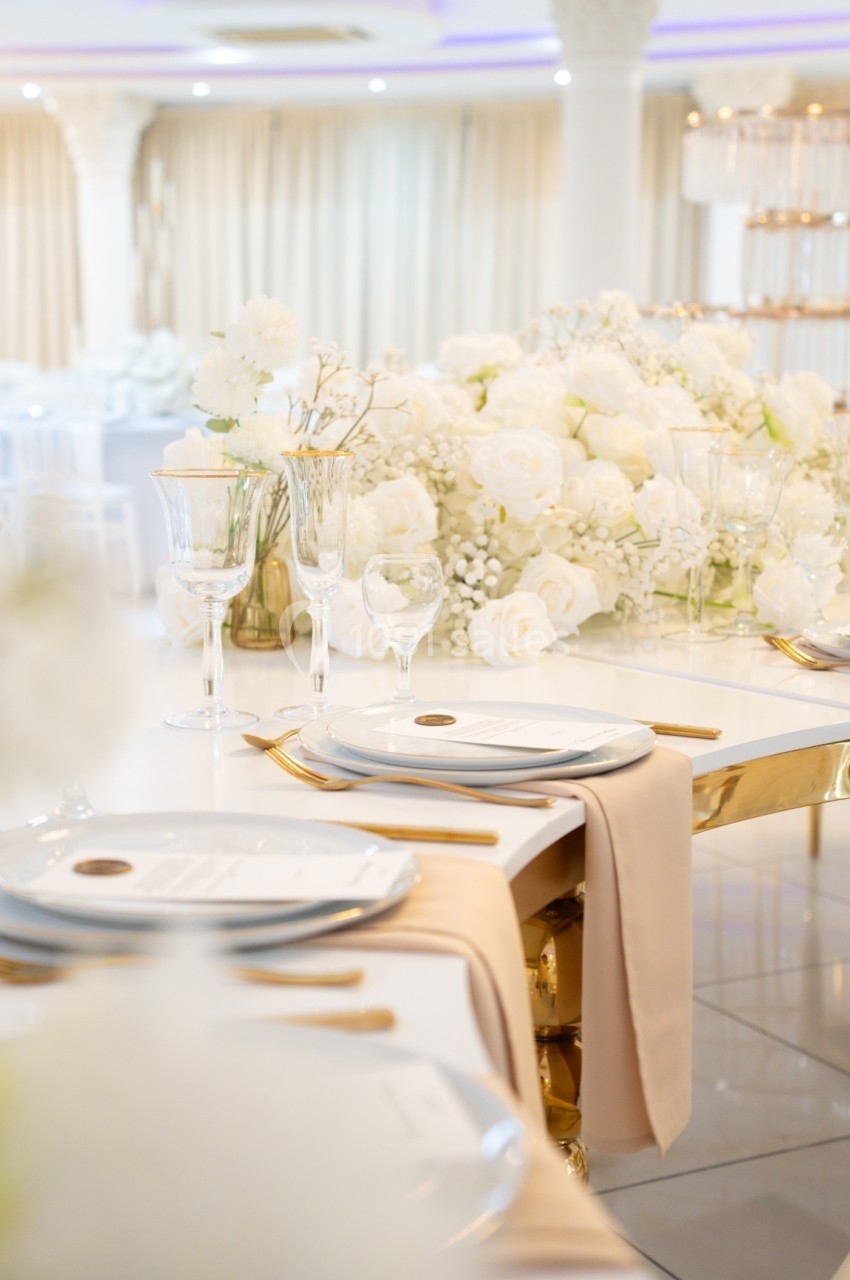 Table élégamment dressée avec vaisselle blanche, nappes beiges et décoration florale blanche dans une salle lumineuse.