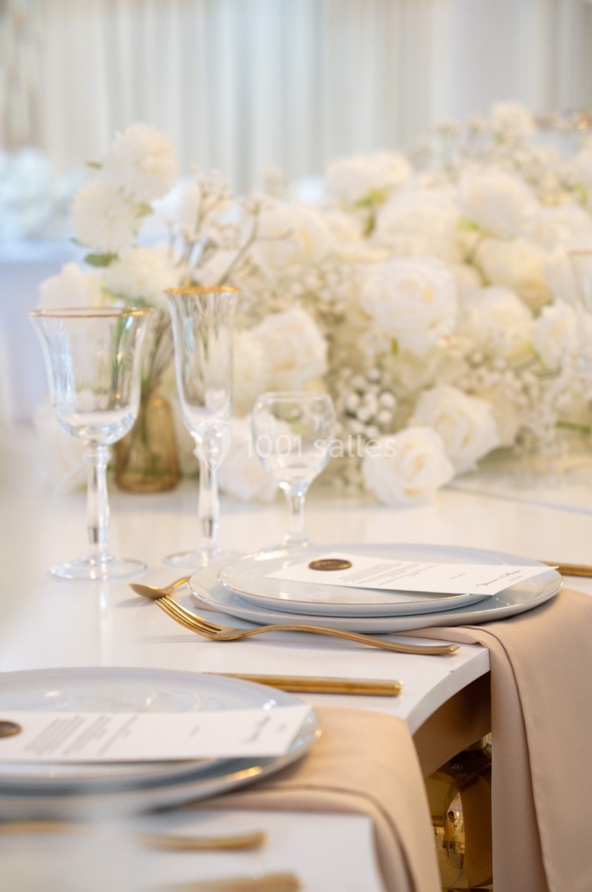 Table élégamment dressée avec assiettes blanches, couverts dorés et décoration florale blanche en arrière-plan.