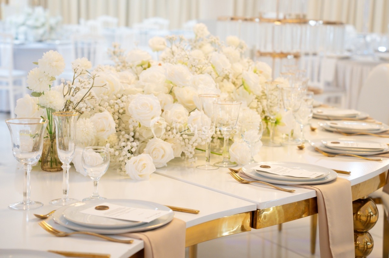 Table élégamment dressée avec nappes blanches, vaisselle raffinée et décoration florale blanche abondante.