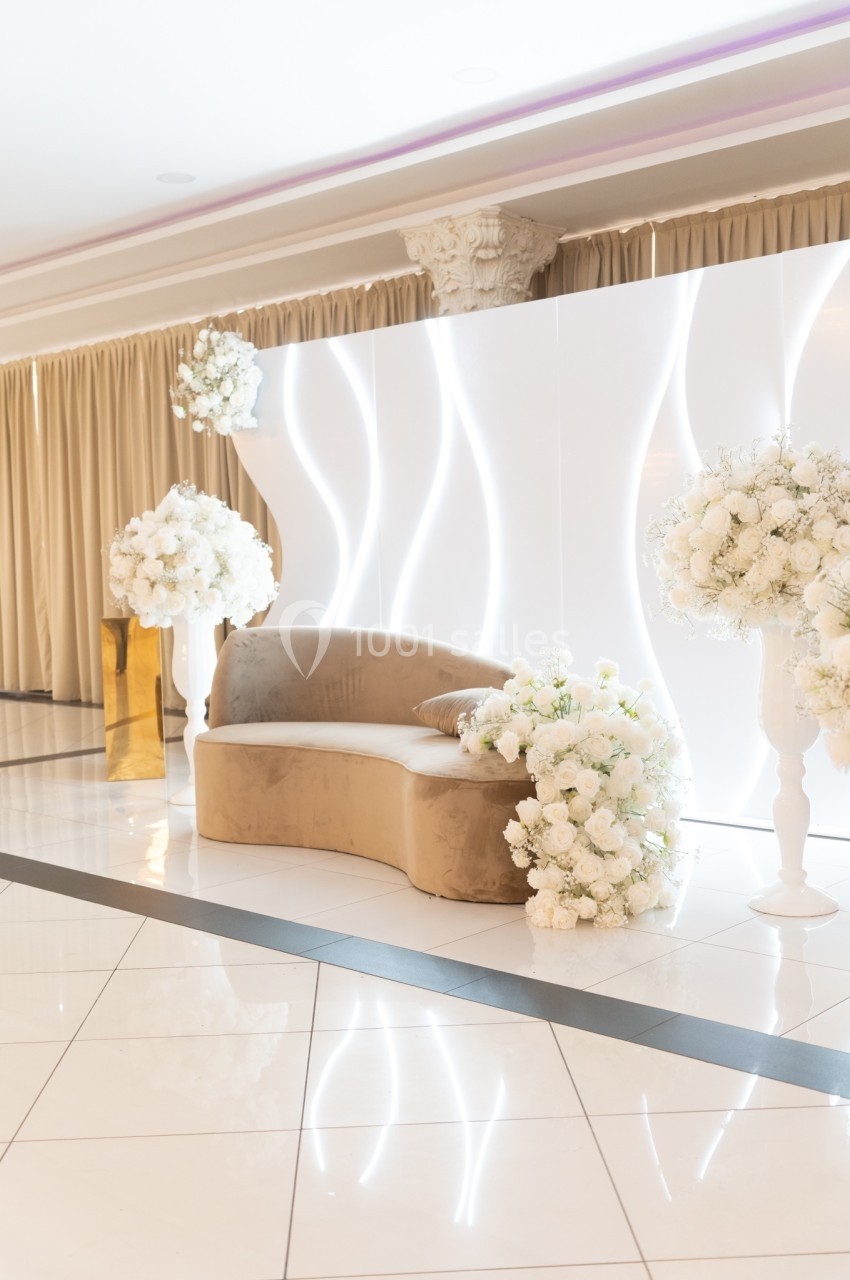 Canapé beige entouré de compositions florales blanches devant un mur lumineux aux motifs ondulés.