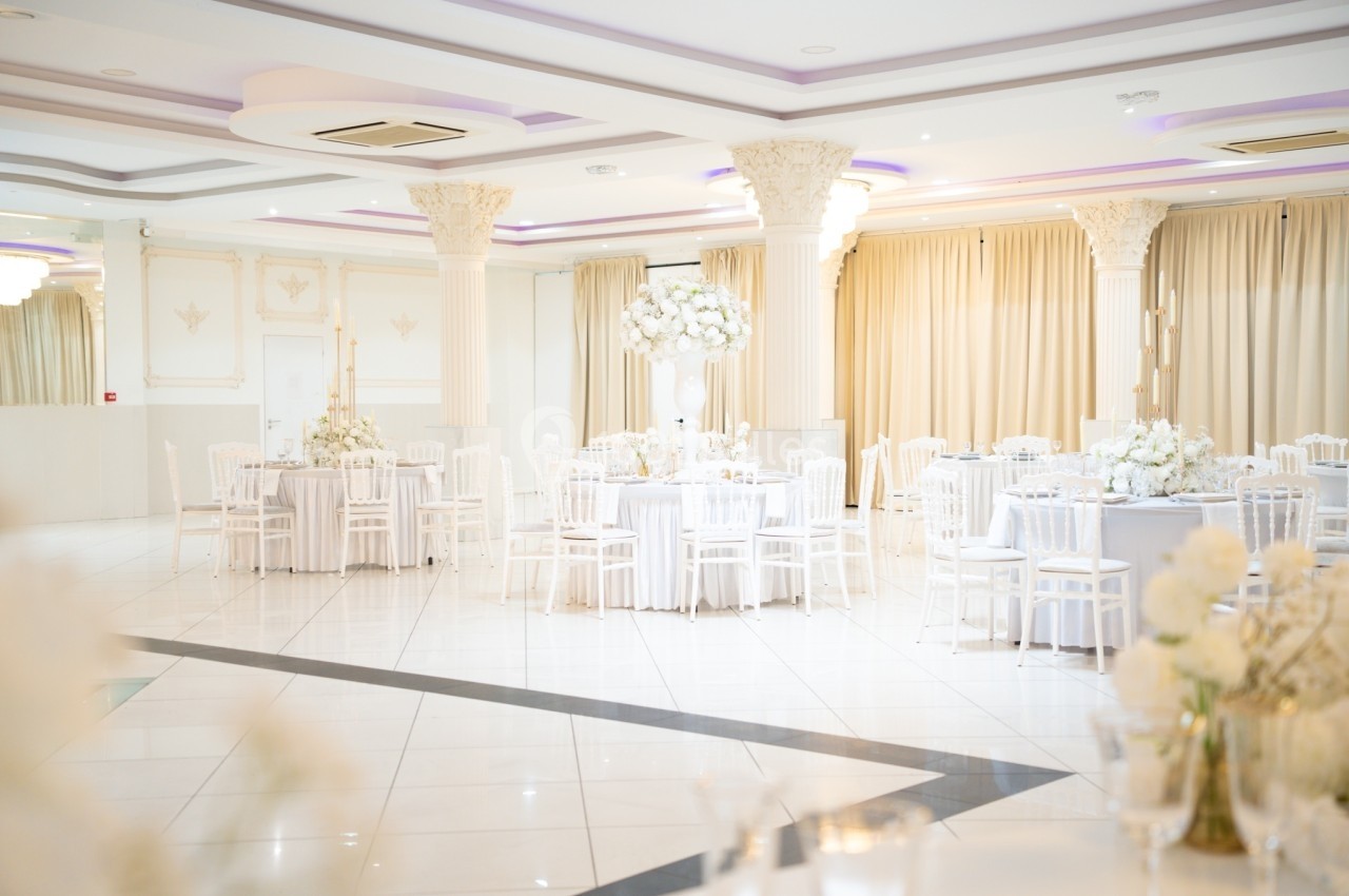 Salle de réception lumineuse avec tables décorées de fleurs blanches, chaises blanches et rideaux beiges.