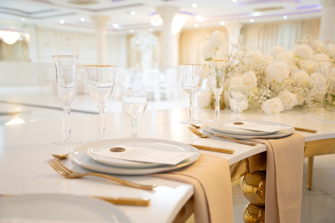 Table élégamment dressée avec nappes beige, vaisselle blanche, couverts dorés et décorations florales blanches.