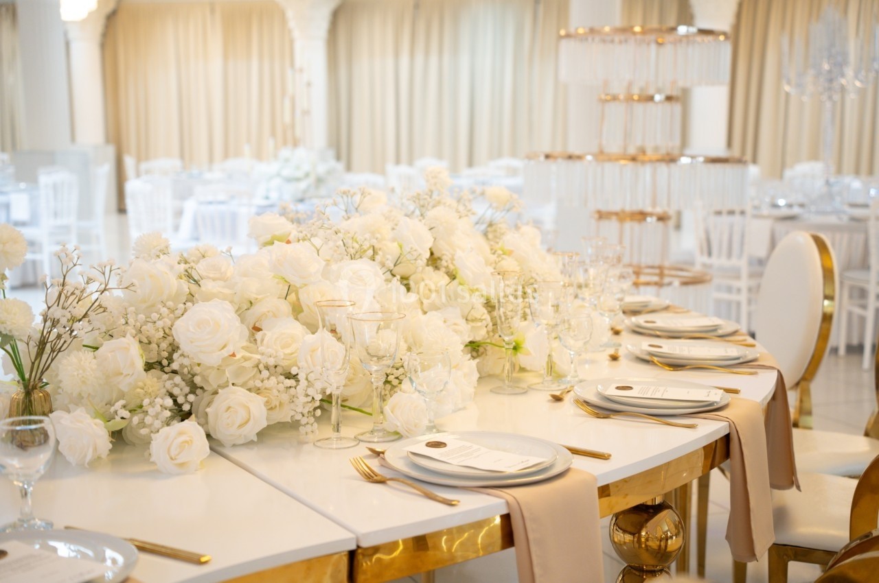 Table élégante décorée de roses blanches, nappes dorées et vaisselle raffinée dans une salle de réception lumineuse.
