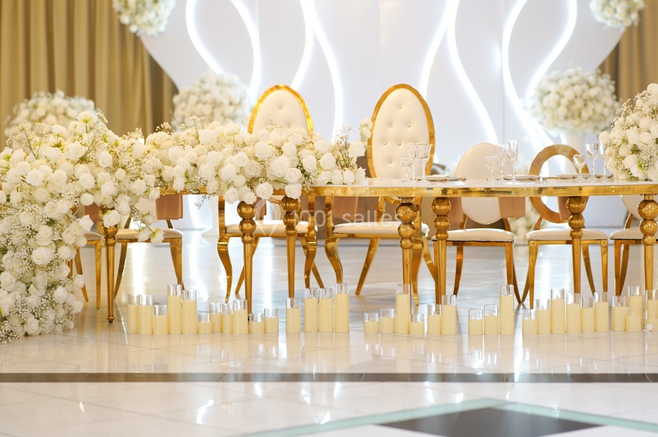 Table décorée avec des chaises dorées, ornée de fleurs blanches et entourée de bougies dans une salle élégante.
