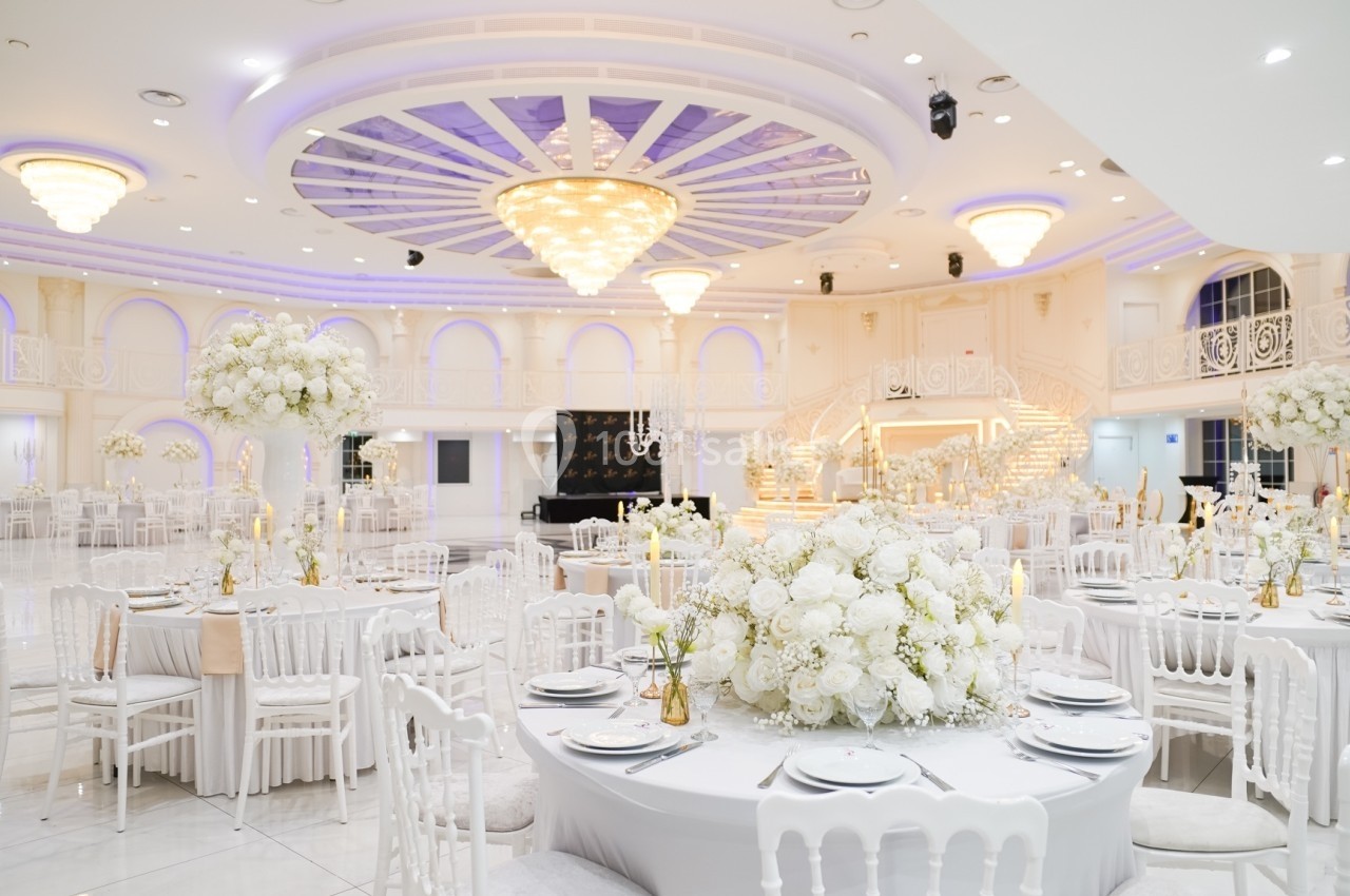 Salle de réception élégante avec tables décorées de fleurs blanches, chaises blanches et grand lustre central.