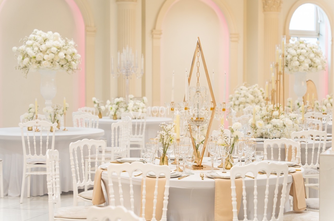 Salle de réception élégante avec tables décorées de fleurs blanches, chaises blanches et lumière douce.