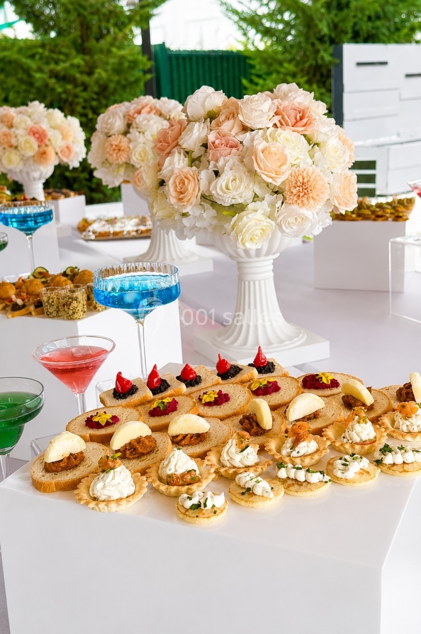 Plateau de canapés variés disposés sur une table blanche, entouré de cocktails colorés et de bouquets de fleurs.