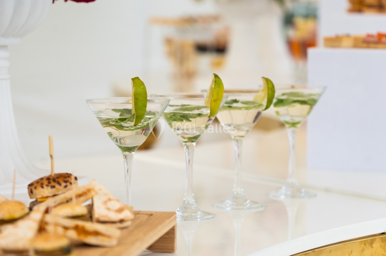 Quatre verres de cocktail garnis de feuilles de menthe et de tranches de citron vert, posés sur une table blanche.