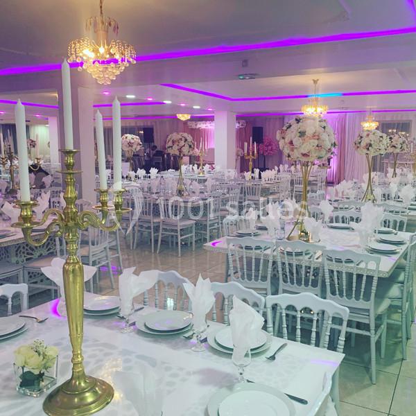 Salle de réception décorée avec des tables dressées, nappes blanches, chandeliers dorés et éclairage rose.