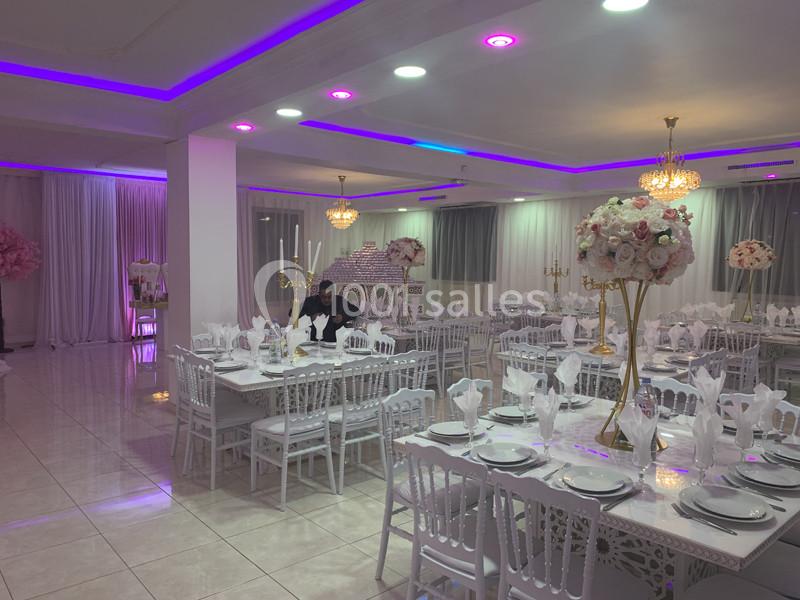 Salle de réception élégante avec tables décorées de nappes blanches, centres de table fleuris et éclairage violet.