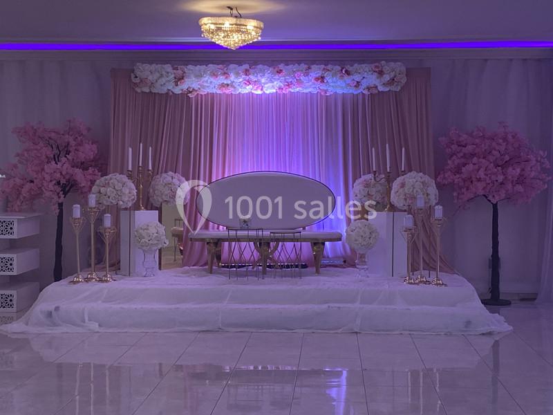 Décor de scène avec canapé ovale, rideaux roses, fleurs blanches et roses, éclairage violet et chandeliers dorés.