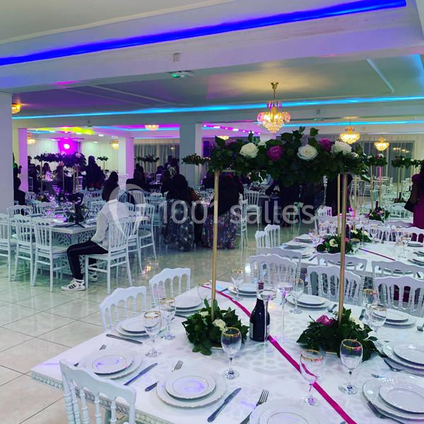 Salle de réception décorée avec des tables dressées, des chaises blanches et des arrangements floraux, éclairage coloré.