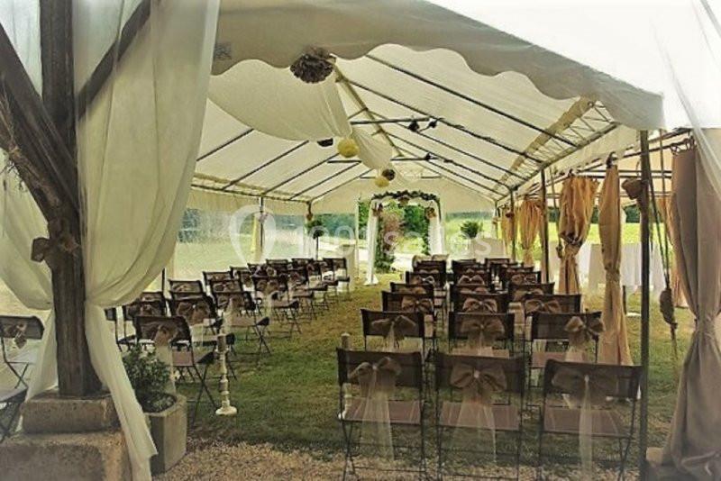 Chaises décorées alignées sous une tente blanche pour une cérémonie en extérieur, avec une arche florale au fond.