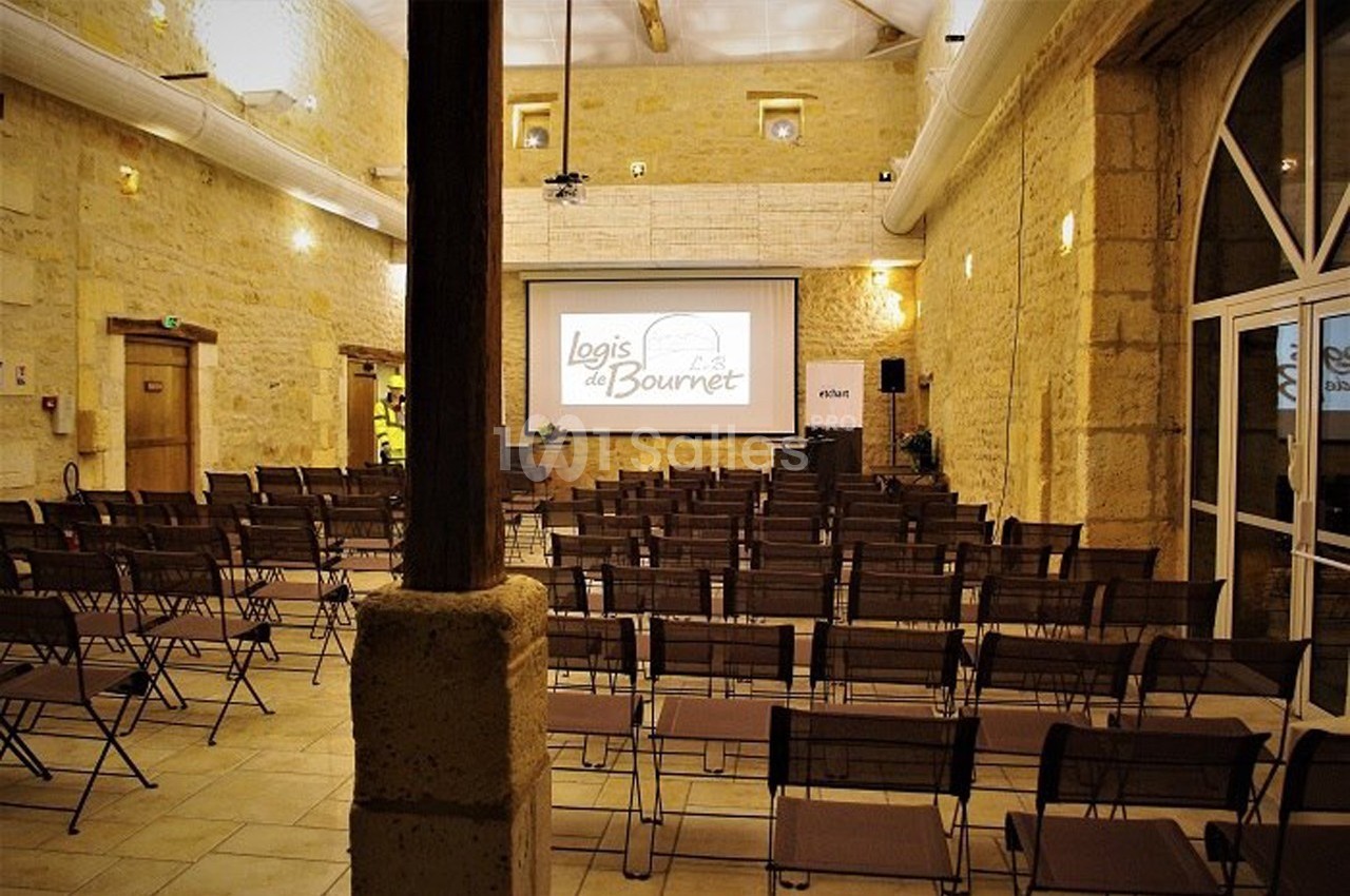 Salle de conférence en pierre avec des rangées de chaises pliantes et un écran de projection allumé au fond.