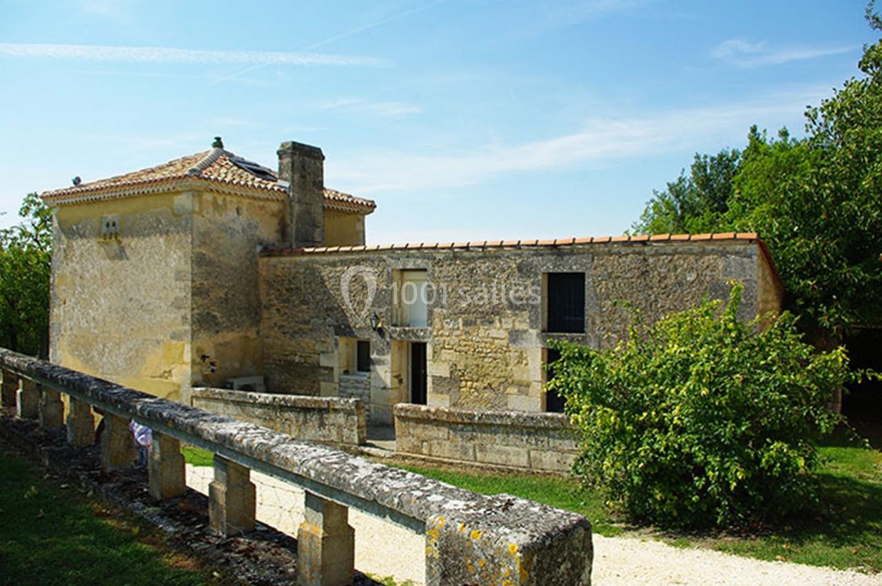 Bâtisse en pierre ancienne avec toit en tuiles, entourée de verdure et d'un muret en pierre sous un ciel dégagé.