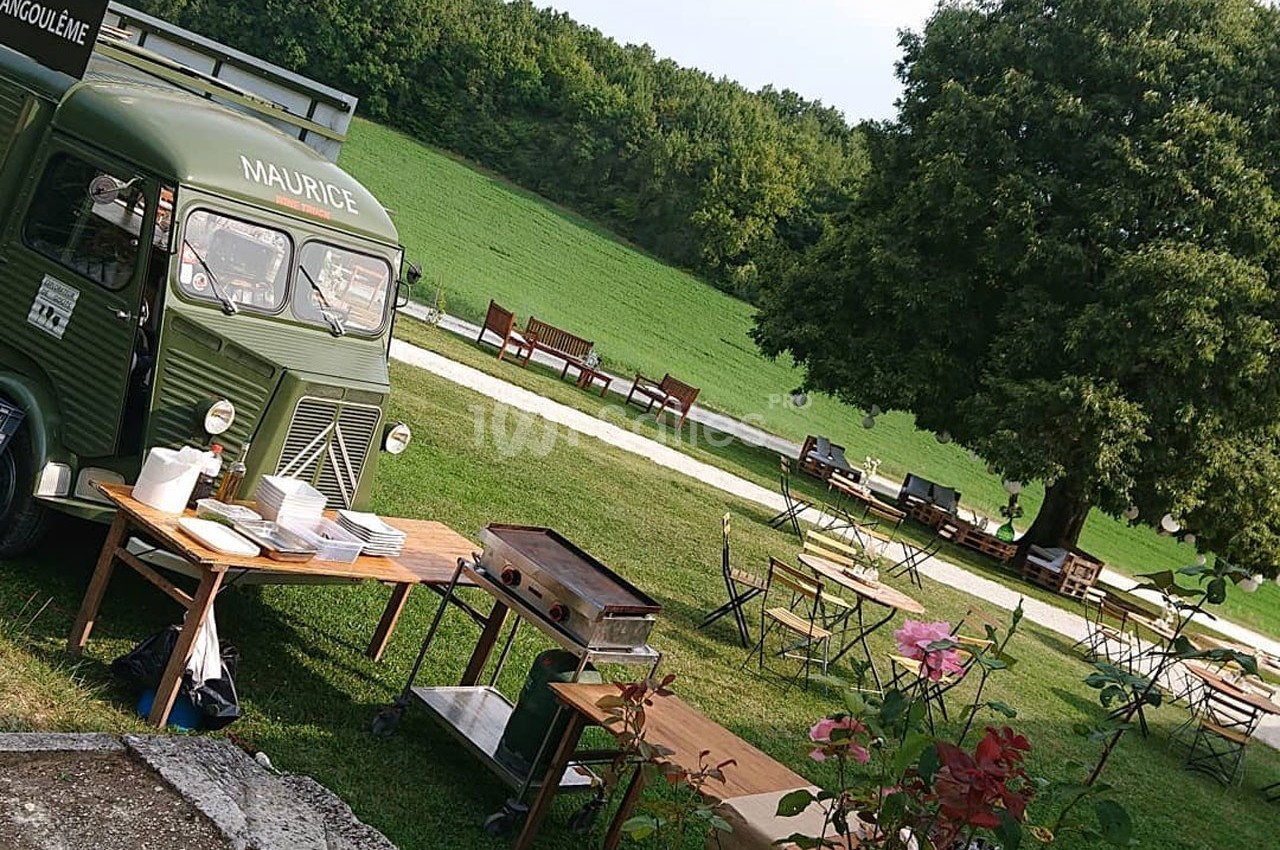 Camion-restaurant vert stationné dans un parc avec tables en bois, chaises et végétation environnante.