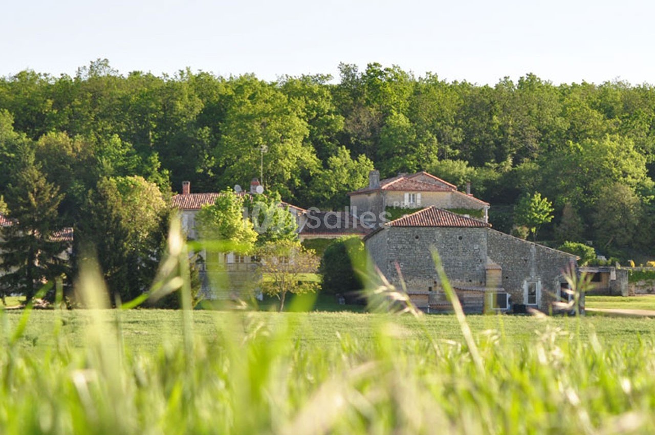 Vue d'un bâtiment en pierre entouré de verdure et d'arbres, avec un champ herbeux au premier plan.