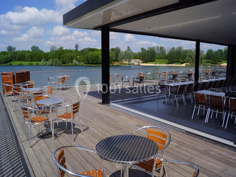 Terrasse en bois avec tables et chaises en métal, offrant une vue sur un lac entouré de verdure par temps ensoleillé.