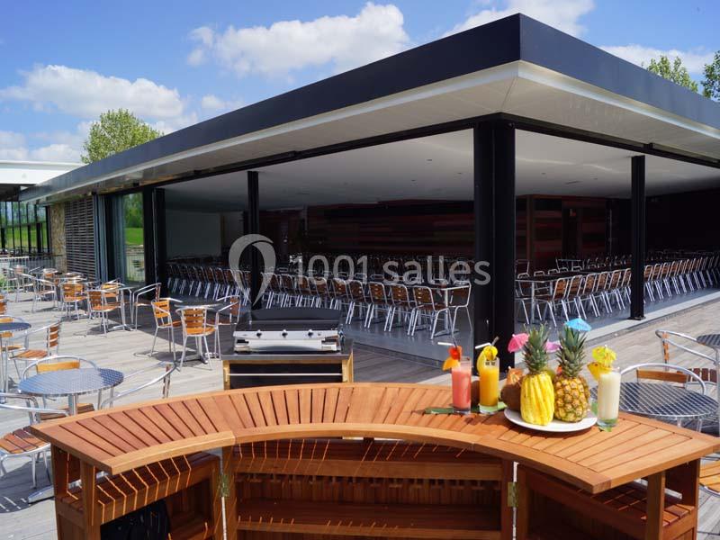Terrasse avec mobilier en métal, bar en bois au premier plan et salle intérieure avec chaises alignées en arrière-plan.