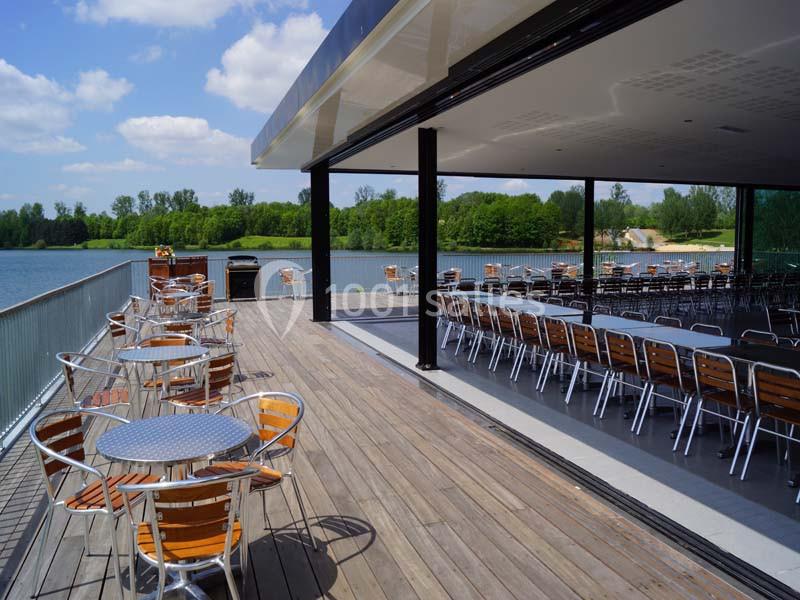 Terrasse en bois avec tables et chaises en métal, donnant sur un lac entouré de verdure par une journée ensoleillée.