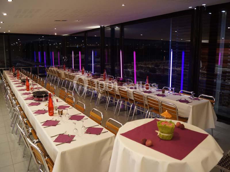Salle de réception avec longues tables dressées, nappes blanches, serviettes violettes et éclairage coloré en arrière-plan.